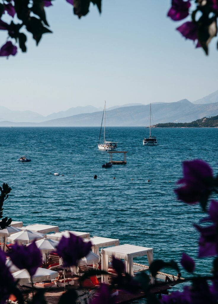 Serene view of sailboats on the clear waters of Ksamil, Albania, with beach cabanas and purple flowers in the foreground—highlighted in every Ksamil Travel Guide.